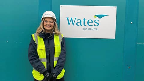 Alice is wearing a hi vis jacket and hard hat. She is smiling in front of a turquoise background with a white sign which has the Wates Residential logo on it.
